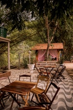 Wooden tables and chairs are arranged on a patterned patio under a dense canopy of trees. In the background, a quaint structure with a red-tiled roof and pink curtains adds a charming touch. Vibrant greenery surrounds the area, giving it a lush, natural feel.