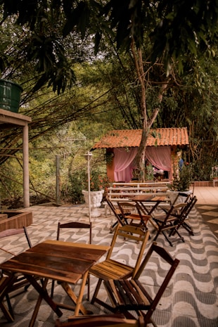 Wooden tables and chairs are arranged on a patterned patio under a dense canopy of trees. In the background, a quaint structure with a red-tiled roof and pink curtains adds a charming touch. Vibrant greenery surrounds the area, giving it a lush, natural feel.