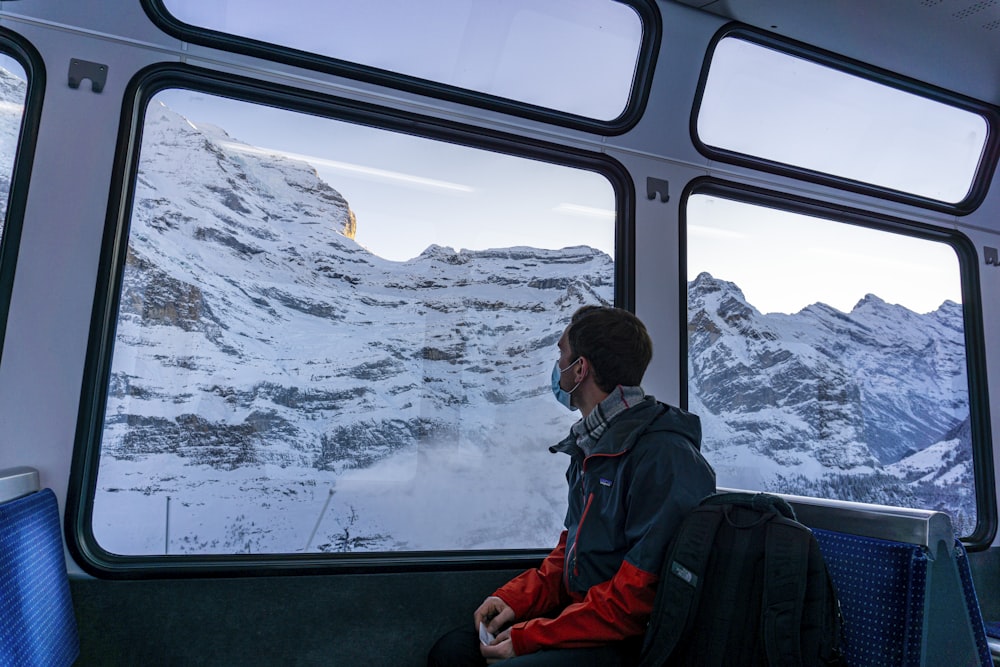 man in black jacket and red backpack sitting on train window looking at snow covered mountain