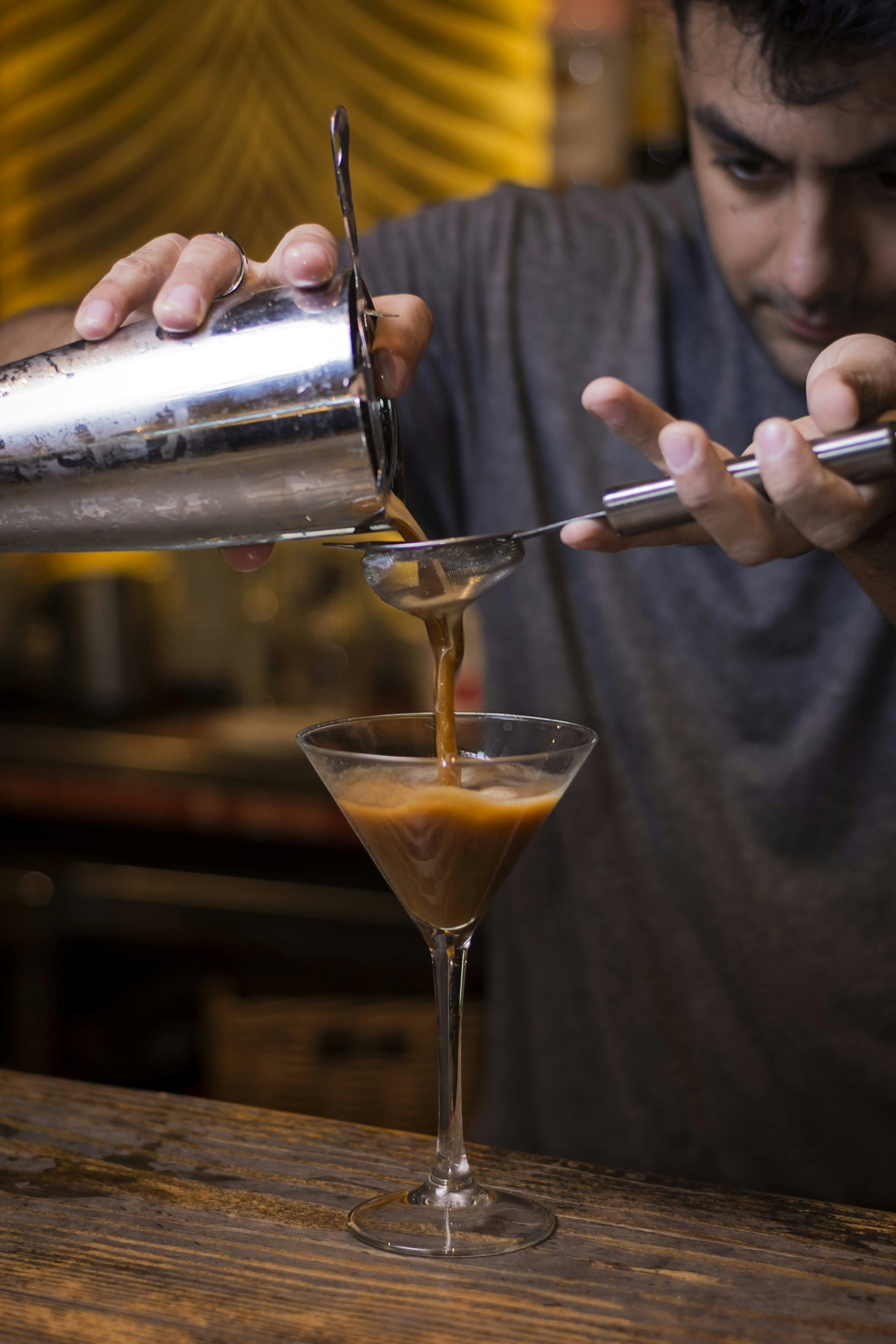 man in gray crew neck shirt holding stainless steel cocktail shaker pouring orange liquid on clear