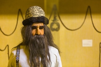 A figure with a long dark beard and a hat inscribed with calligraphy, wearing a traditional garment with decorative patterns on the straps. The background features a simple beige wall with abstract, curved black line drawings and a small plaque.