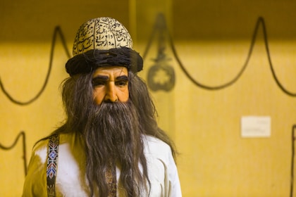 A figure with a long dark beard and a hat inscribed with calligraphy, wearing a traditional garment with decorative patterns on the straps. The background features a simple beige wall with abstract, curved black line drawings and a small plaque.