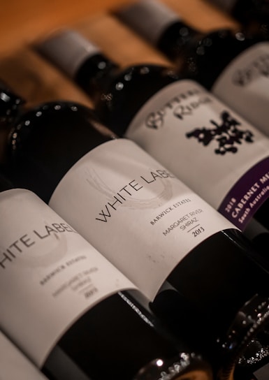 Several wine bottles are arranged in a diagonal line on a wooden surface. The labels are prominently displayed with various names and design elements, while the caps are a metallic silver.