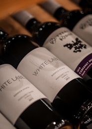 Several wine bottles are arranged in a diagonal line on a wooden surface. The labels are prominently displayed with various names and design elements, while the caps are a metallic silver.