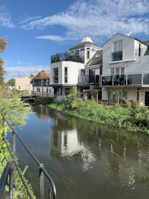 A picturesque scene alongside a calm, flowing river featuring modern houses with balconies and large windows. Several of the houses are white with glass railings. The buildings are mirrored in the reflective water of the river. The banks are lush with green foliage, and the sky is mostly clear with some scattered clouds. A metal railing runs along the sidewalk next to the river, leading towards a distant bridge crossing the water.