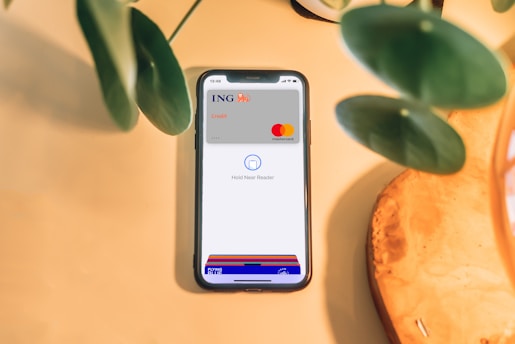 A smartphone is placed on a light wooden surface, displaying a digital wallet interface with an ING credit card and Mastercard logo. Green plant leaves partially frame the top and left sides of the image.