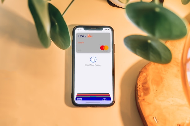 A smartphone is placed on a light wooden surface, displaying a digital wallet interface with an ING credit card and Mastercard logo. Green plant leaves partially frame the top and left sides of the image.