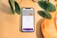 A smartphone is placed on a light wooden surface, displaying a digital wallet interface with an ING credit card and Mastercard logo. Green plant leaves partially frame the top and left sides of the image.