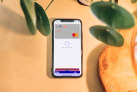 A smartphone is placed on a light wooden surface, displaying a digital wallet interface with an ING credit card and Mastercard logo. Green plant leaves partially frame the top and left sides of the image.