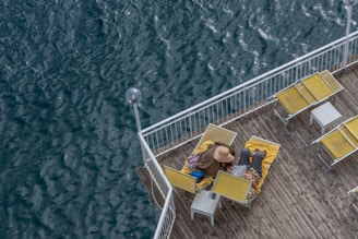 A person wearing a hat and a coat is lying on a yellow lounger on a wooden deck overlooking a body of water. The deck is equipped with multiple sun loungers and small tables, arranged in a tidy manner. The railing encloses the area providing safety from the adjacent water.