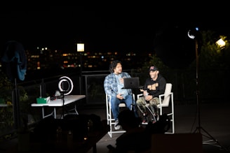 Two people are seated on a rooftop at night, equipped with video conferencing equipment. The cityscape in the background is out of focus, suggesting an urban environment. There are several studio lights, a ring light, and a table with electronic devices. The individuals appear to be engaged in a relaxed conversation, possibly part of a podcast or livestream.