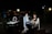 Two people are seated on a rooftop at night, equipped with video conferencing equipment. The cityscape in the background is out of focus, suggesting an urban environment. There are several studio lights, a ring light, and a table with electronic devices. The individuals appear to be engaged in a relaxed conversation, possibly part of a podcast or livestream.