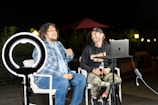 Two people are sitting on chairs outdoors in a nighttime setting, engaged in what appears to be a lively conversation. A ring light is positioned in front of them, providing illumination. One person is wearing a plaid shirt and jeans, while the other is dressed in a hoodie and camouflage pants. A laptop is set up on a nearby table, and a microphone is positioned nearby. There is subtle lighting in the background, including a red umbrella and some outdoor lamps.