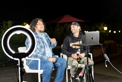 Two people are sitting on chairs outdoors in a nighttime setting, engaged in what appears to be a lively conversation. A ring light is positioned in front of them, providing illumination. One person is wearing a plaid shirt and jeans, while the other is dressed in a hoodie and camouflage pants. A laptop is set up on a nearby table, and a microphone is positioned nearby. There is subtle lighting in the background, including a red umbrella and some outdoor lamps.