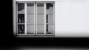 A rectangular window with six glass panes and a white frame is set against a white wall. Subtle reflections and shadows are visible on the glass, indicating an interior environment beyond the window. The window is neatly aligned with the structure, and the surrounding area is mostly plain, giving it a minimalist appearance.