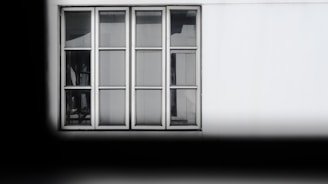 A rectangular window with six glass panes and a white frame is set against a white wall. Subtle reflections and shadows are visible on the glass, indicating an interior environment beyond the window. The window is neatly aligned with the structure, and the surrounding area is mostly plain, giving it a minimalist appearance.