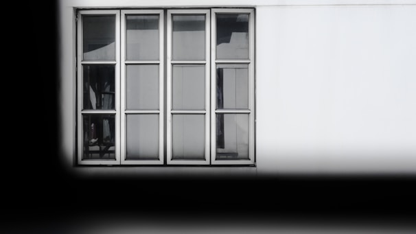 A rectangular window with six glass panes and a white frame is set against a white wall. Subtle reflections and shadows are visible on the glass, indicating an interior environment beyond the window. The window is neatly aligned with the structure, and the surrounding area is mostly plain, giving it a minimalist appearance.