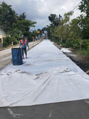 A road under construction is covered with a white tarp. A person wearing a safety vest and a helmet is standing near a blue barrel on the left side. The road is flanked by trees and some houses are visible on the left. The sky is cloudy, contributing to a calm atmosphere.