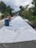 A road under construction is covered with a white tarp. A person wearing a safety vest and a helmet is standing near a blue barrel on the left side. The road is flanked by trees and some houses are visible on the left. The sky is cloudy, contributing to a calm atmosphere.