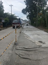 A road under construction is flanked by trees and power lines. The surface is partially covered with large sheets, and a truck is parked in the left lane with a person standing on it.