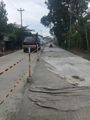 A road under construction is flanked by trees and power lines. The surface is partially covered with large sheets, and a truck is parked in the left lane with a person standing on it.