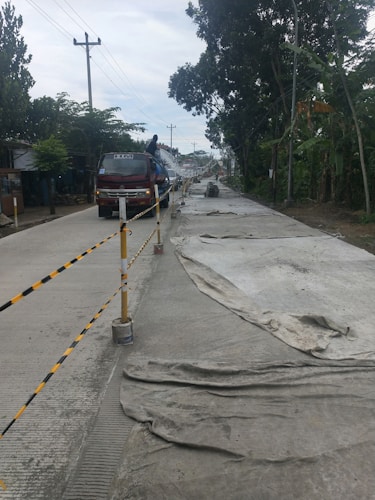 A road under construction is flanked by trees and power lines. The surface is partially covered with large sheets, and a truck is parked in the left lane with a person standing on it.