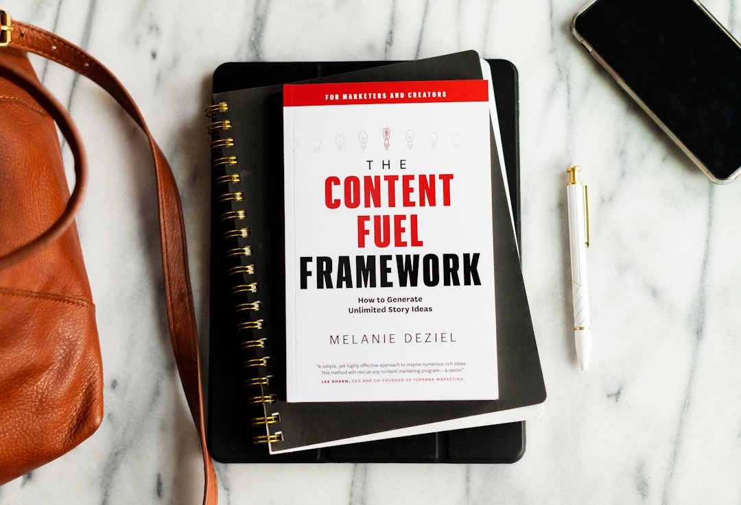 the content fuel framework book next to a purse, A flatlay of a workspace, with a white marble background. Includes a book, black notebook and planner, a fancy white pen, smartphone/iphone, and brown leather bag.