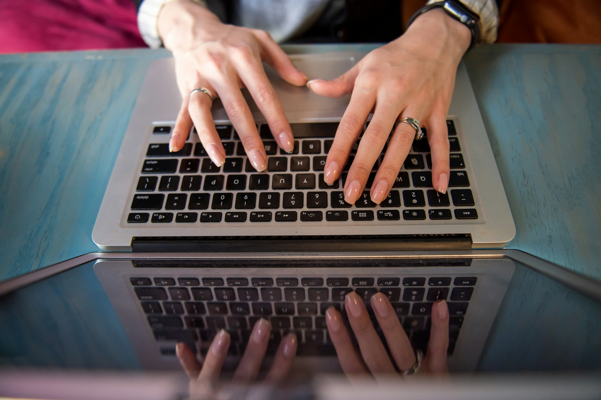 Woman's manicured hands typing on an Apple keyboard. She is wearing silver rings and a smart watch / apple watch. 