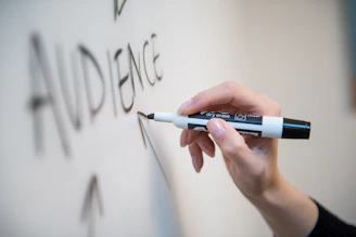 A hand holding a black dry erase marker writing on a white board