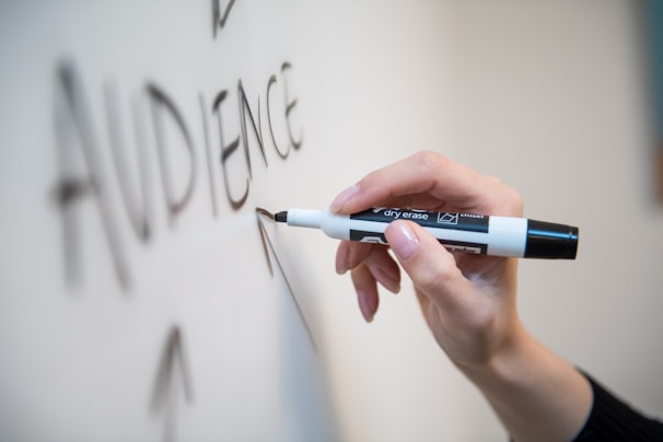 A hand is holding a black dry erase marker and writing the word 'AUDIENCE' on a whiteboard.