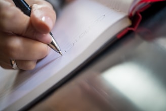 A close-up view of a person's hand writing with a pen in a notebook. The notebook is open, revealing lined pages, and the hand is wearing a ring.