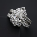 A close-up view of an elegant diamond ring featuring a large, pear-shaped central diamond surrounded by a halo of smaller round diamonds. The ring band is also adorned with diamonds, enhancing its luxurious appearance. The dark background contrasts with the brilliance of the diamonds, highlighting their sparkle.