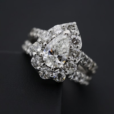 A close-up view of an elegant diamond ring featuring a large, pear-shaped central diamond surrounded by a halo of smaller round diamonds. The ring band is also adorned with diamonds, enhancing its luxurious appearance. The dark background contrasts with the brilliance of the diamonds, highlighting their sparkle.