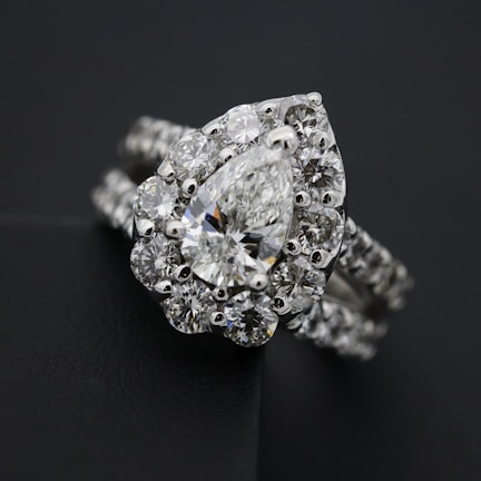 A close-up view of an elegant diamond ring featuring a large, pear-shaped central diamond surrounded by a halo of smaller round diamonds. The ring band is also adorned with diamonds, enhancing its luxurious appearance. The dark background contrasts with the brilliance of the diamonds, highlighting their sparkle.