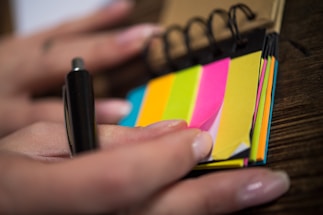 A close-up of a hand holding a small spiral notepad with colorful sticky notes and a pen. The sticky notes are in bright pink, yellow, green, orange, and blue, and the hand is preparing to use one.