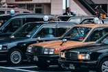 A group of traditional Japanese taxis is parked in a row, featuring their characteristic roof signs. The cars include black and yellow models. Two individuals are standing next to the taxis, likely drivers in formal attire.