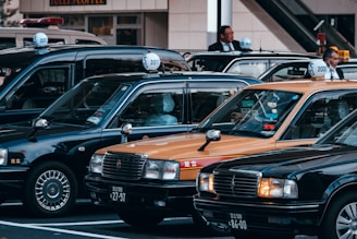 A group of traditional Japanese taxis is parked in a row, featuring their characteristic roof signs. The cars include black and yellow models. Two individuals are standing next to the taxis, likely drivers in formal attire.