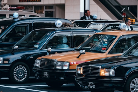 A group of traditional Japanese taxis is parked in a row, featuring their characteristic roof signs. The cars include black and yellow models. Two individuals are standing next to the taxis, likely drivers in formal attire.