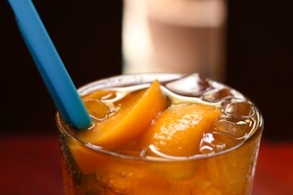 A glass filled with iced tea and cubes of peach slices, with a blue straw protruding from the top. The beverage appears cold and refreshing, with the peach slices glistening in the light.