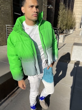 A man is standing outdoors, wearing a vibrant green and gray puffer jacket and white pants with 'Choices' written on them. The sunlight casts a shadow on the concrete pavement. Behind him, there are steps and a building with brick walls.