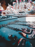 A table displays various jewelry pieces, including chains and necklaces, with a blurred background full of colorful items in bags. The lighting creates a reflective effect on the metallic surfaces.