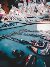 A table displays various jewelry pieces, including chains and necklaces, with a blurred background full of colorful items in bags. The lighting creates a reflective effect on the metallic surfaces.