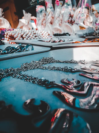 A table displays various jewelry pieces, including chains and necklaces, with a blurred background full of colorful items in bags. The lighting creates a reflective effect on the metallic surfaces.