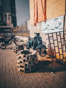 An old scooter is parked against a wall adorned with posters. The scooter is surrounded by a stack of bricks and a metal frame. Several motorbikes are parked along a fence in the background under a clear sky. A large construction site is partially visible in the distance.
