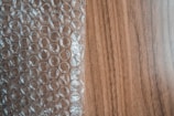 A sheet of transparent bubble wrap lies on a wooden surface, partially covering it. The bubble wrap consists of evenly spaced, raised circular bubbles that create a pattern of repeating circles. The wooden surface has a light brown color with visible grain patterns.
