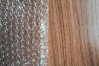 A sheet of transparent bubble wrap lies on a wooden surface, partially covering it. The bubble wrap consists of evenly spaced, raised circular bubbles that create a pattern of repeating circles. The wooden surface has a light brown color with visible grain patterns.