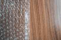 A sheet of transparent bubble wrap lies on a wooden surface, partially covering it. The bubble wrap consists of evenly spaced, raised circular bubbles that create a pattern of repeating circles. The wooden surface has a light brown color with visible grain patterns.