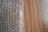 A sheet of transparent bubble wrap lies on a wooden surface, partially covering it. The bubble wrap consists of evenly spaced, raised circular bubbles that create a pattern of repeating circles. The wooden surface has a light brown color with visible grain patterns.