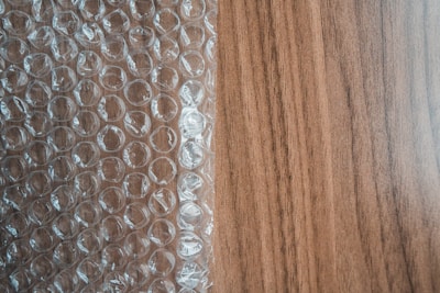 A sheet of transparent bubble wrap lies on a wooden surface, partially covering it. The bubble wrap consists of evenly spaced, raised circular bubbles that create a pattern of repeating circles. The wooden surface has a light brown color with visible grain patterns.