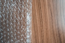 A sheet of transparent bubble wrap lies on a wooden surface, partially covering it. The bubble wrap consists of evenly spaced, raised circular bubbles that create a pattern of repeating circles. The wooden surface has a light brown color with visible grain patterns.